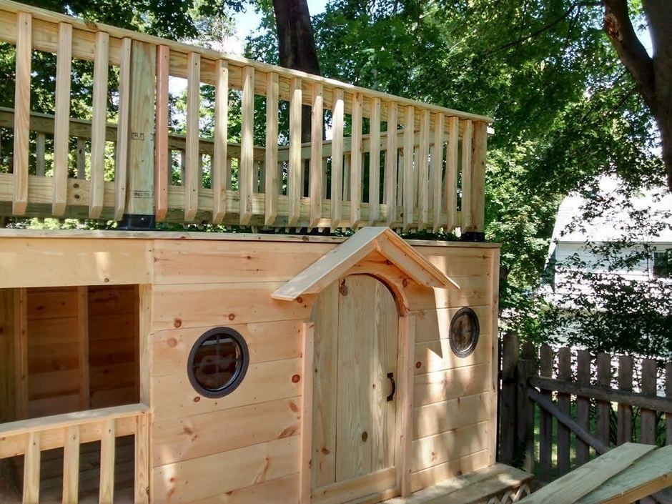 Wooden backyard playhouse with a balcony and round windows, surrounded by trees and a fence.